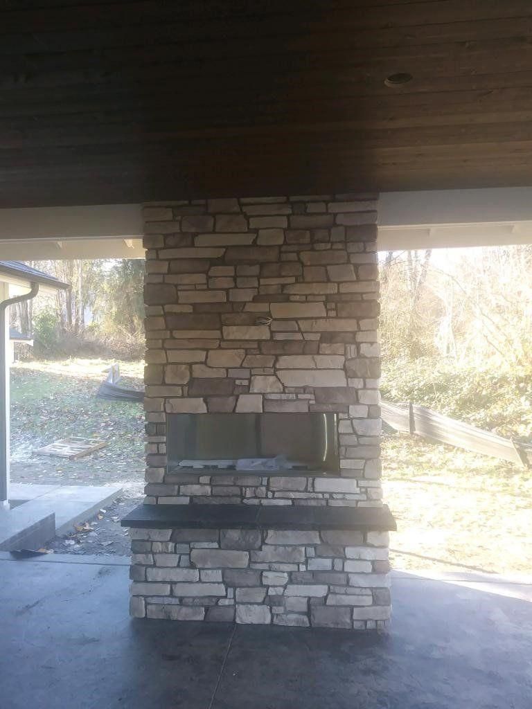 Stone fireplace under a covered outdoor patio with a concrete floor and open yard beyond