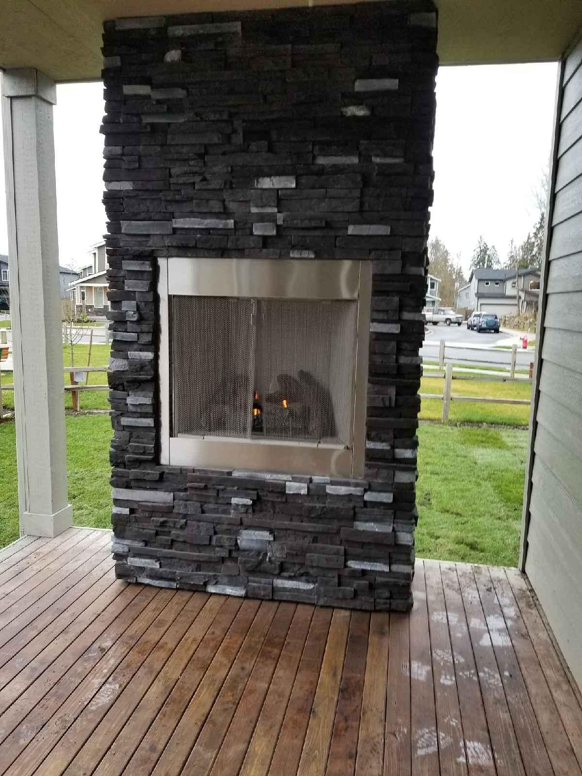 Stone outdoor fireplace on a wooden porch, with grass and a parking lot in the background
