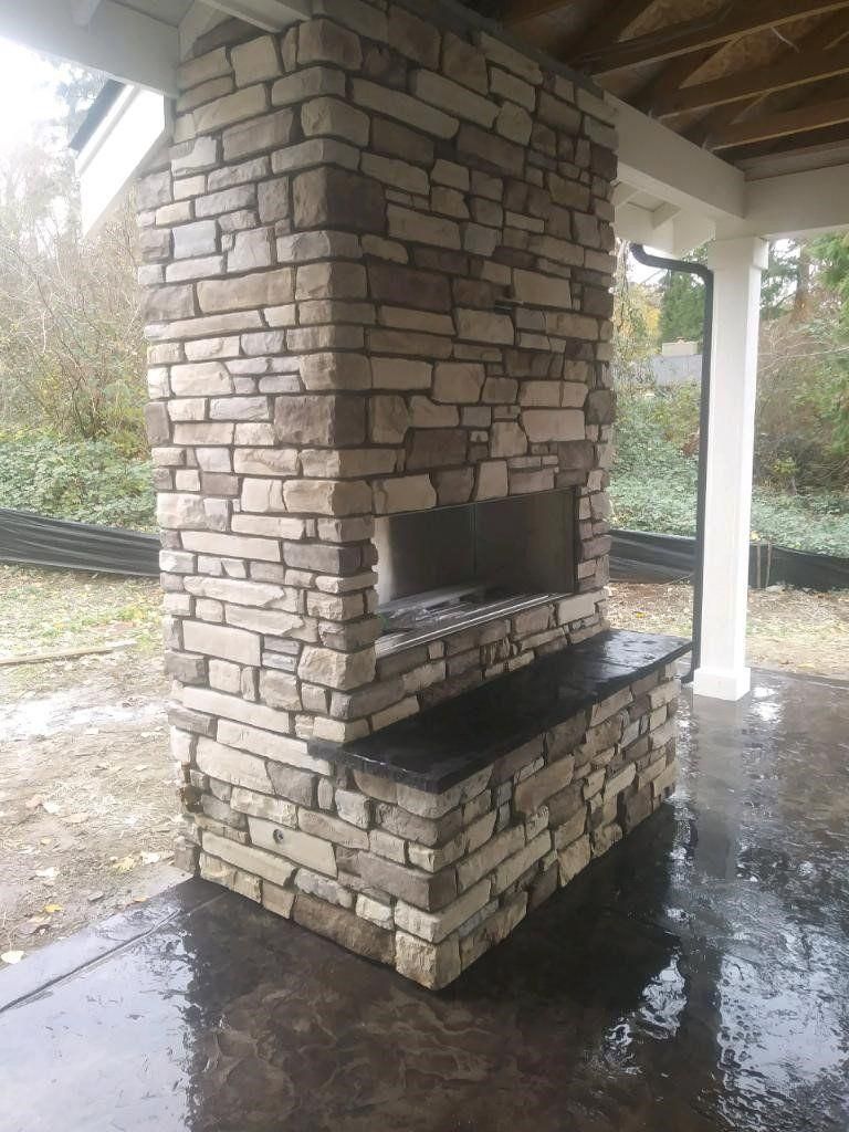 Stone outdoor fireplace under a covered patio, with wet concrete floor and white support columns.