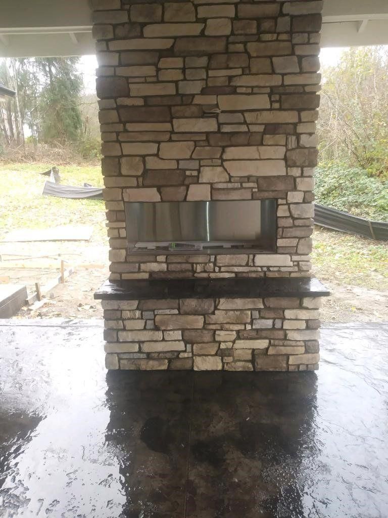 Stone outdoor fireplace on a covered patio with a dark wet floor and grassy yard beyond