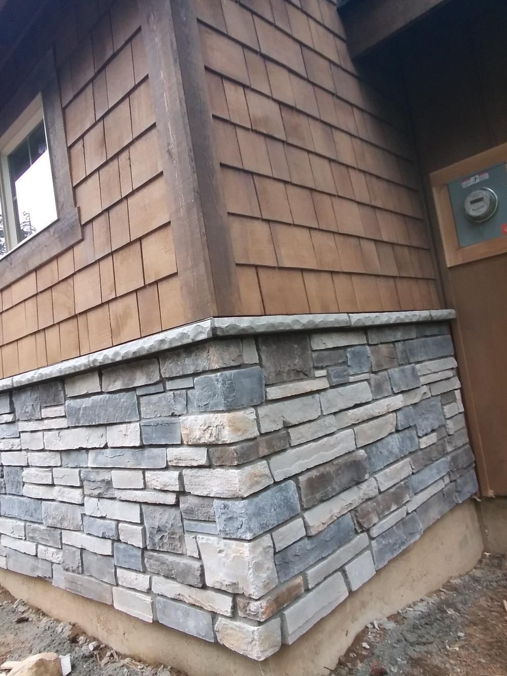 Corner of a house with tan shingle siding and a stacked stone foundation wall рядом to a door.