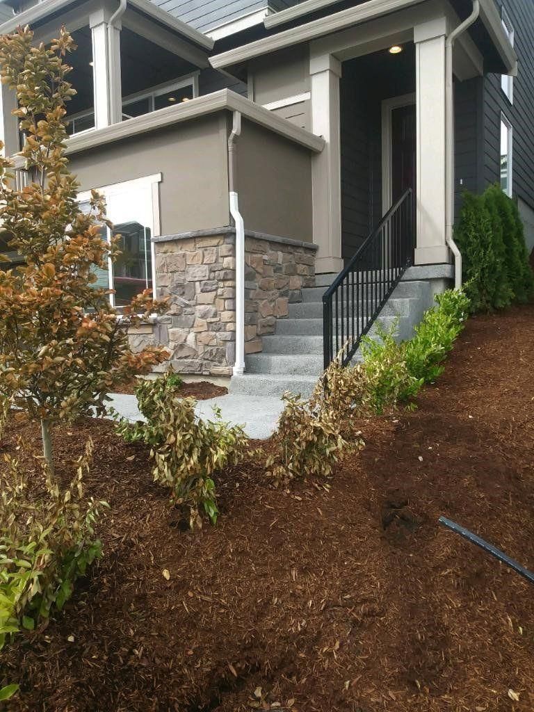 Front steps to a gray house with stone siding, black railing, and a landscaped dirt slope.