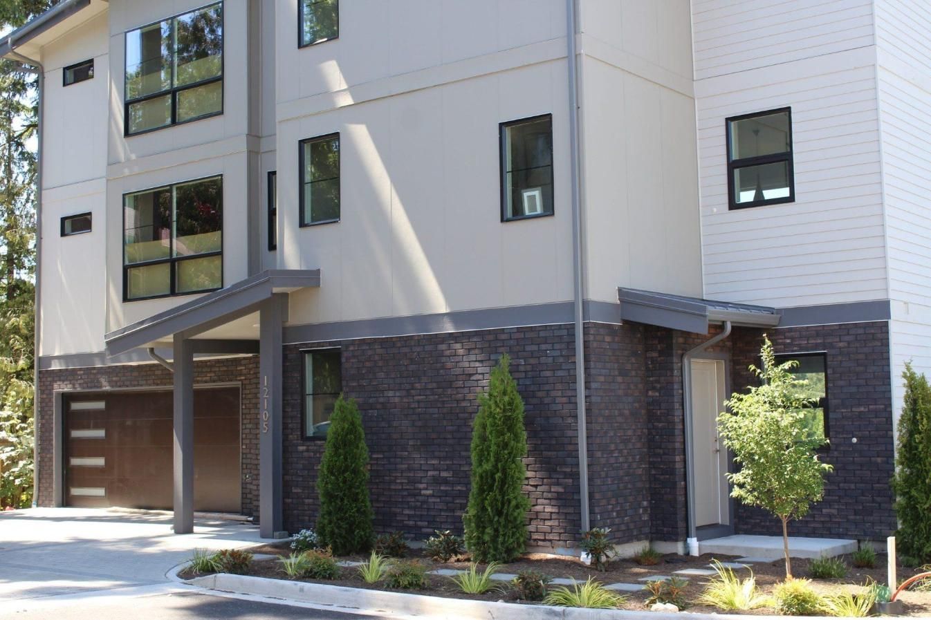 Modern apartment building exterior with garage, gray siding, brick base, and landscaped trees