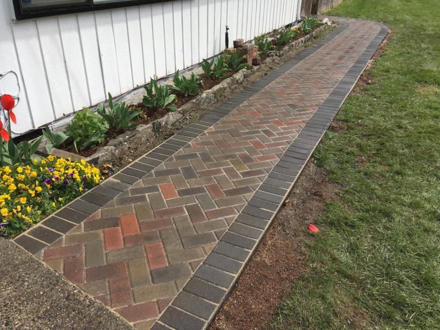 Brick garden path beside a white building, bordered by flower beds and grass.