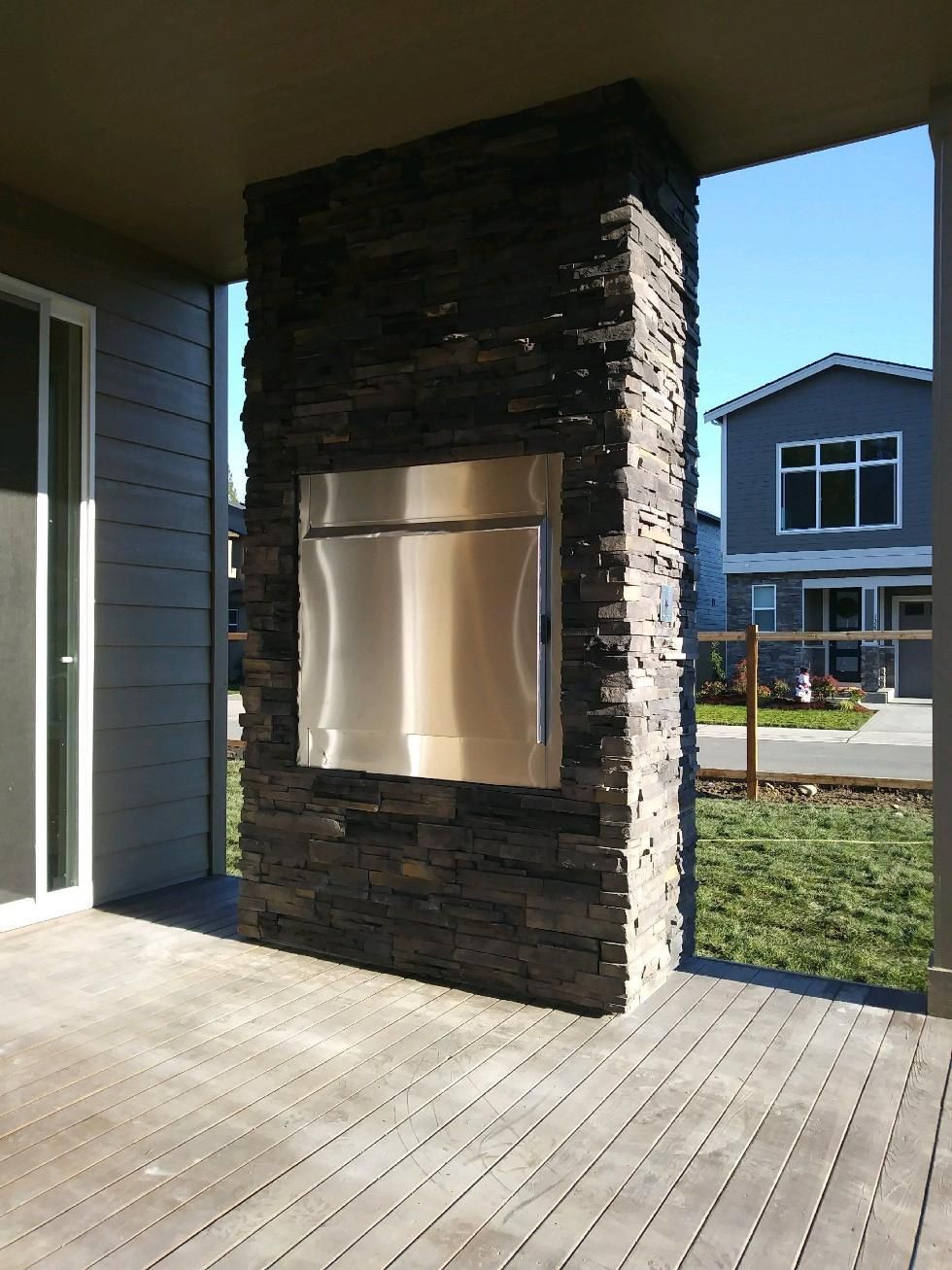 Covered outdoor patio with a stone column, stainless grill, and wooden deck facing a house.