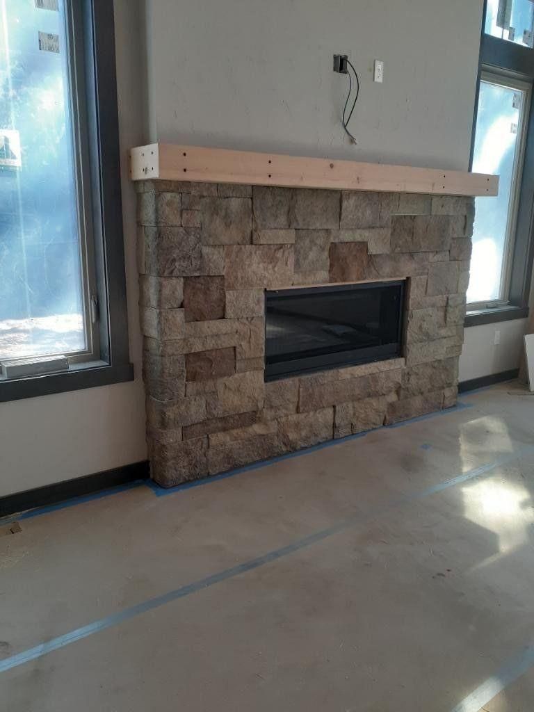 Stone fireplace with a light wood mantel in an empty room beside a window.