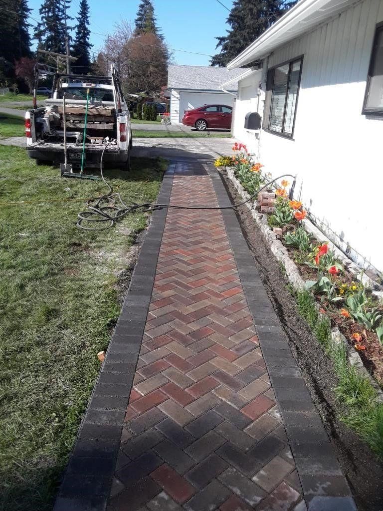 Brick walkway beside a white house and flower bed, with a small trailer parked on the lawn.
