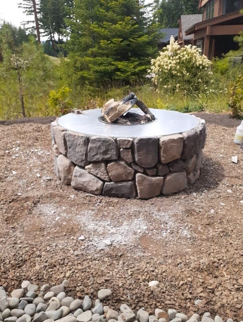 Stone fire pit with wood in the center, set in a gravel yard beside a cabin and trees.
