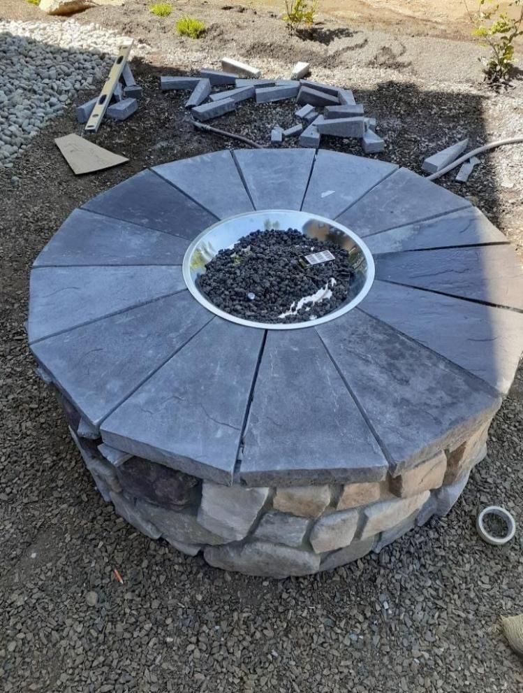 Round stone fire pit with gray paver top and scattered bricks on gravel ground
