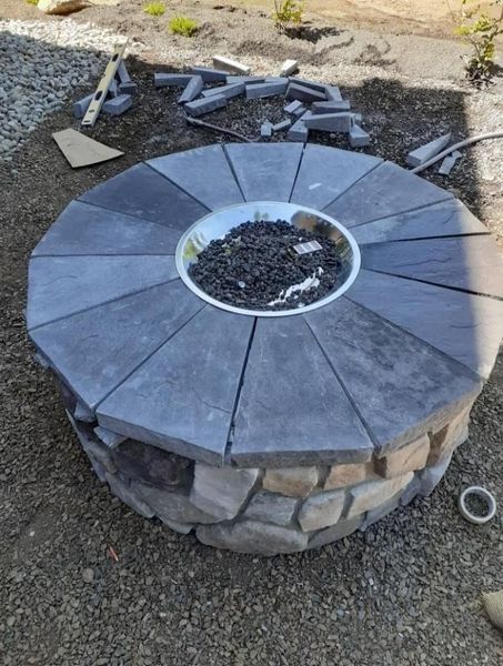 Round stone fire pit with gray paver top and scattered bricks on gravel ground