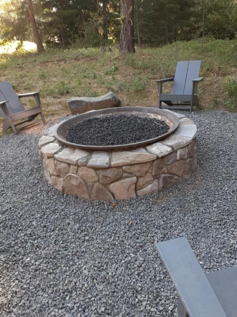 Stone fire pit surrounded by Adirondack chairs on a gravel patio in a wooded yard