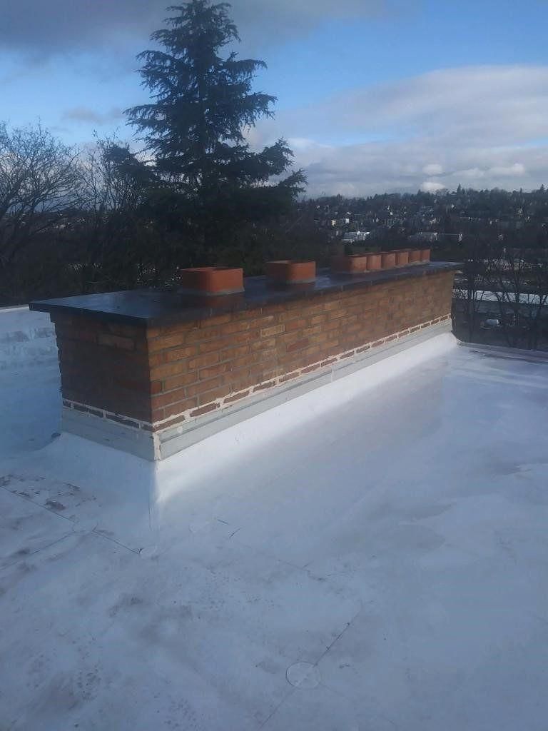 Snow-covered rooftop with a brick chimney structure and trees in the background under a cloudy sky