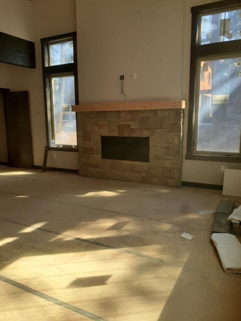 Empty living room with a stone fireplace and tall windows, sunlight on bare carpet.