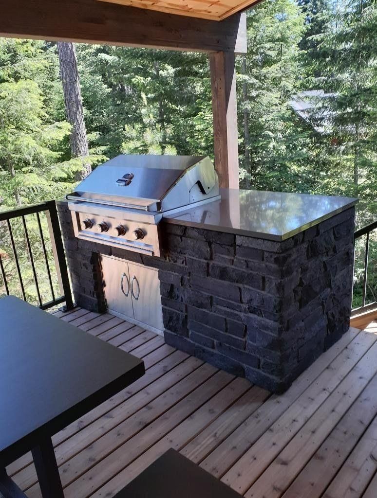 Covered deck with a stainless grill and stone outdoor counter, surrounded by trees.