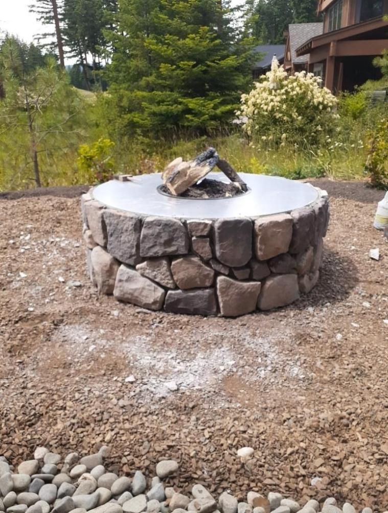 Stone fire pit with a metal ring in a wooded yard, surrounded by gravel and a house deck in the background