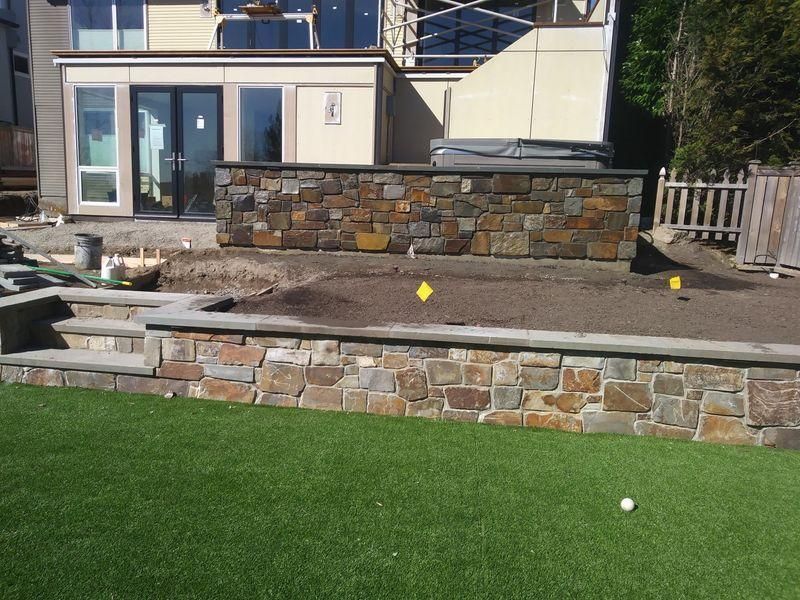 Backyard stone patio with raised retaining wall, steps, and a green lawn in front of a house