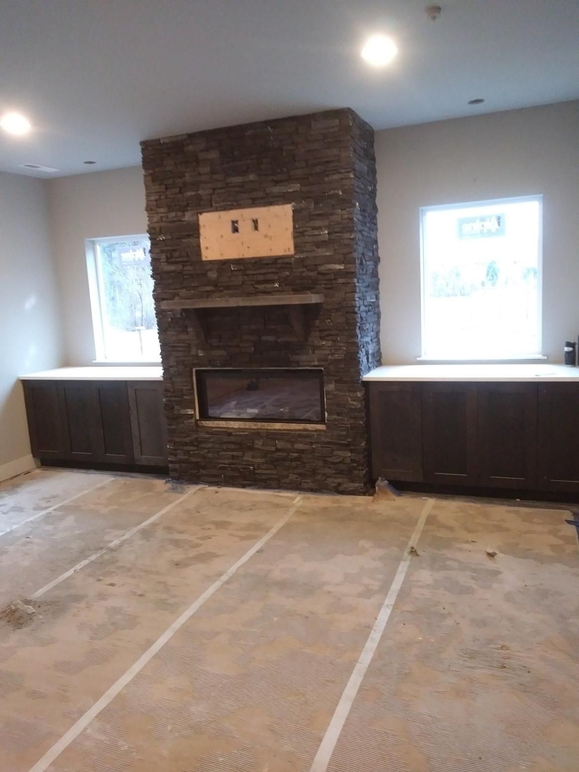 Living room with stone fireplace, two windows, dark built-in cabinets, and unfinished flooring with taped seams