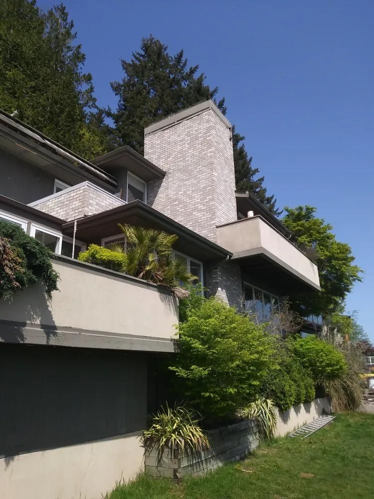 Modern hillside house with white stucco walls, dark roof, and lush greenery under a blue sky