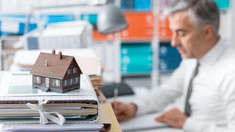 A man is sitting at a desk with a model house on top of a pile of binders.