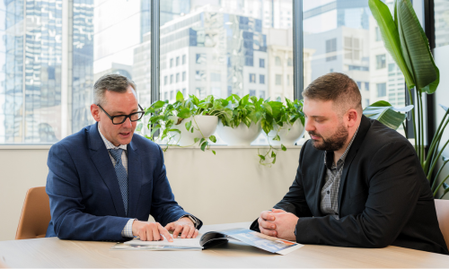 Two men are sitting at a table looking at a book.