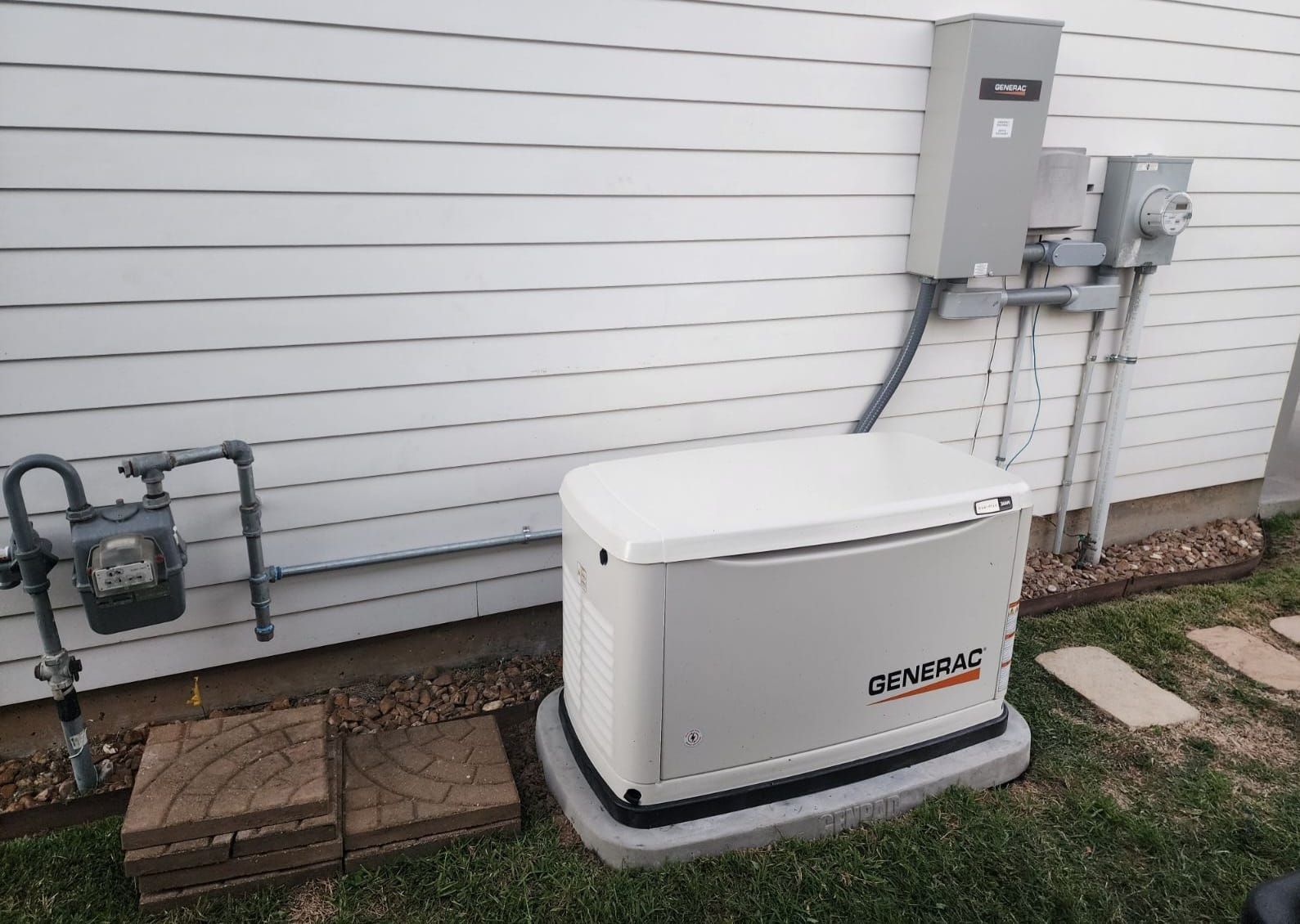 White whole-house generator beside a house’s exterior wall with electrical boxes and piping
