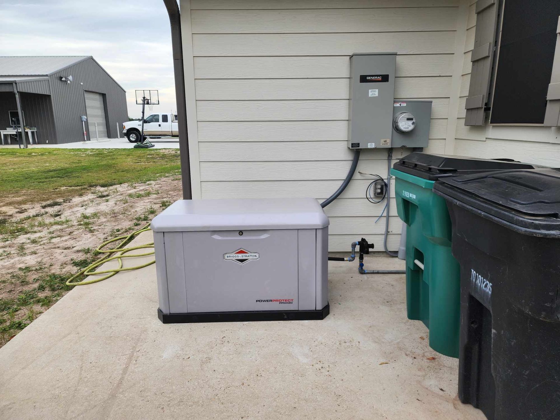 Outdoor generator beside a house wall, with trash bins and utility meter nearby.