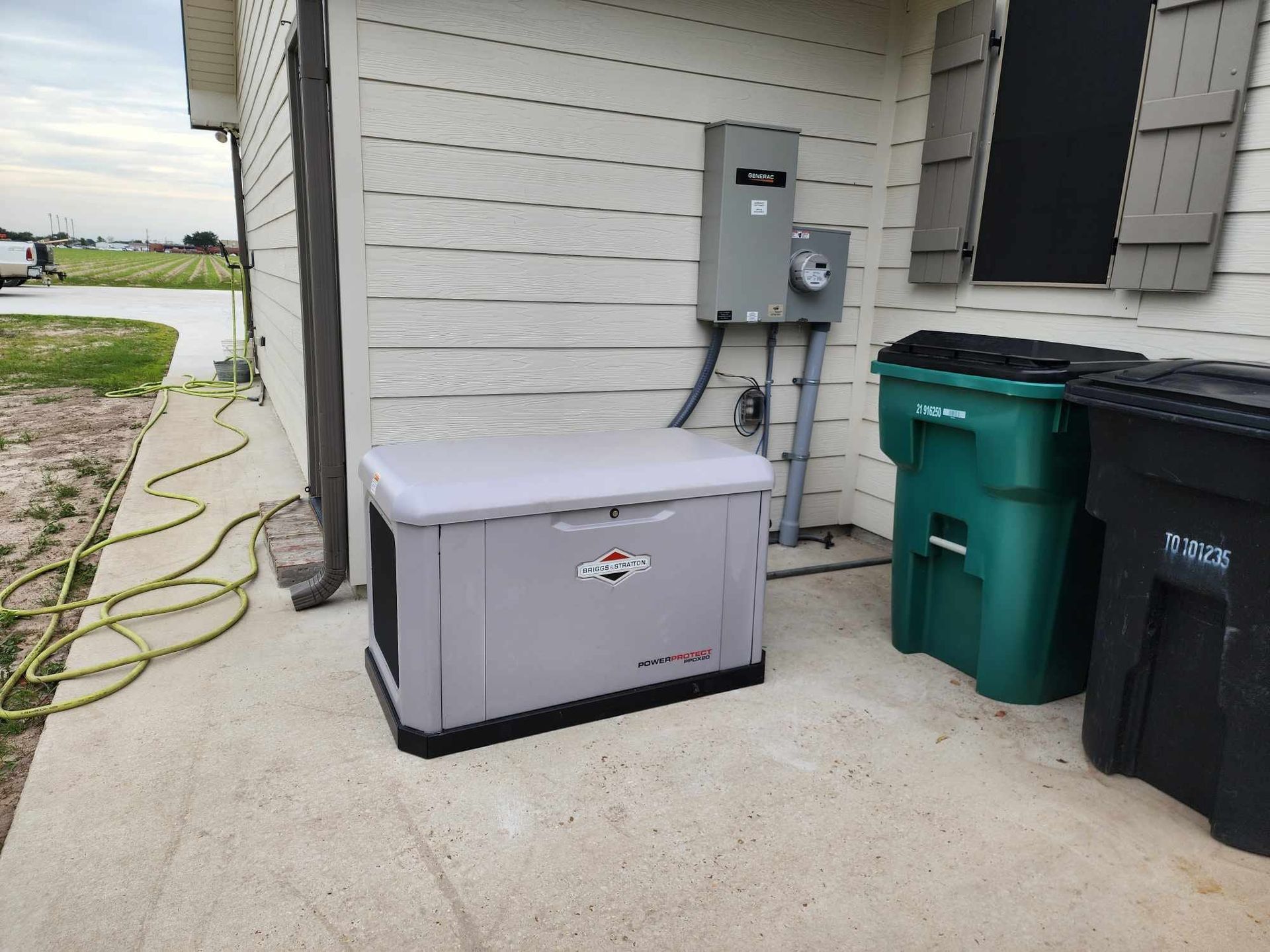 Gray generator beside house wall next to electrical meter and trash bins on a concrete slab