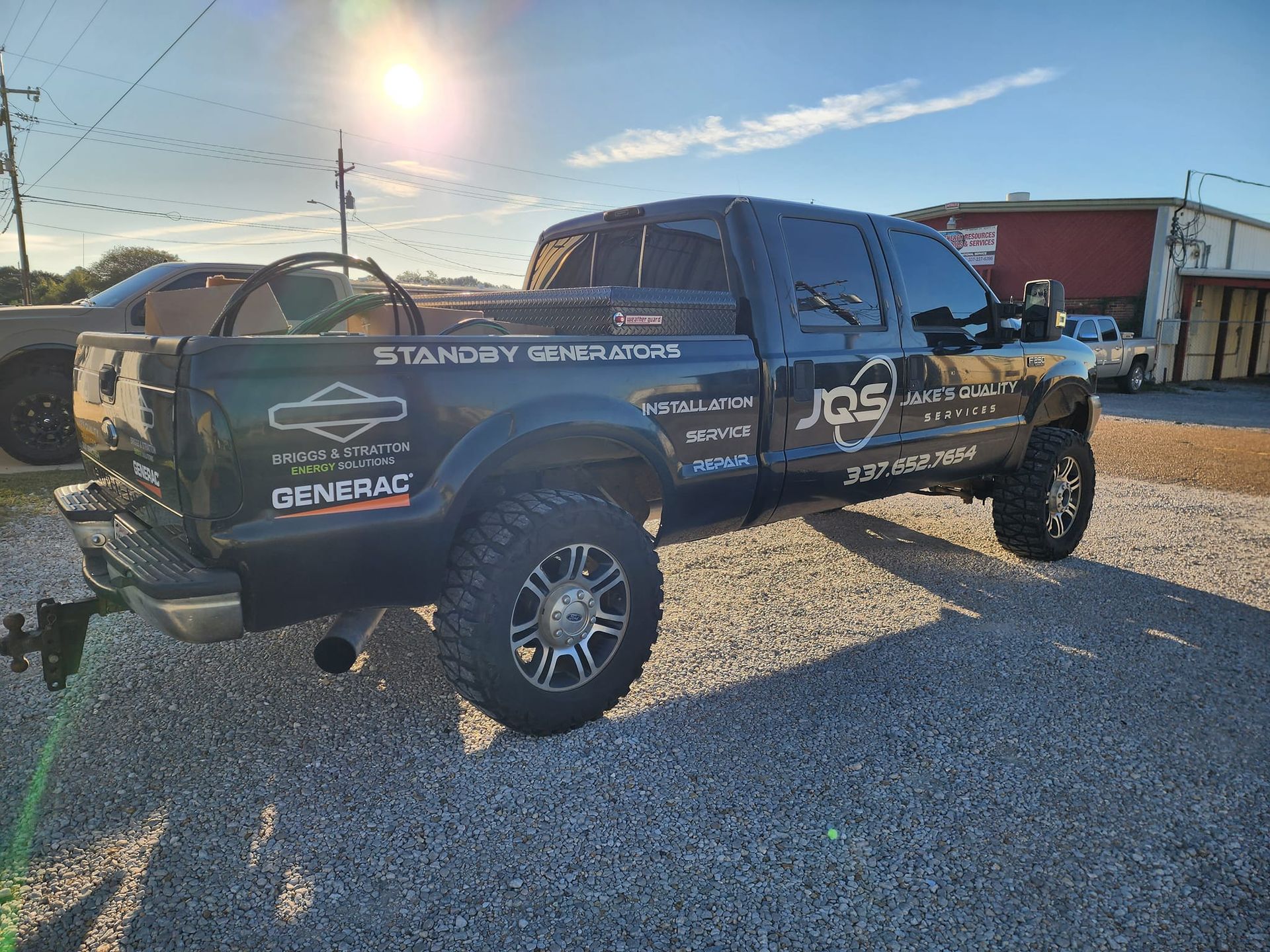 Lifted black pickup truck parked on gravel at sunset, with sponsor decals and off-road tires.