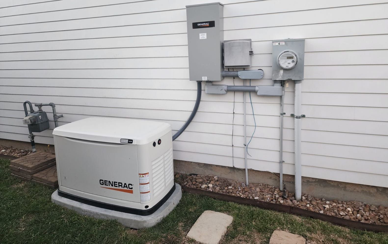 Outdoor generator beside a house wall, connected to gray electrical boxes and conduit.