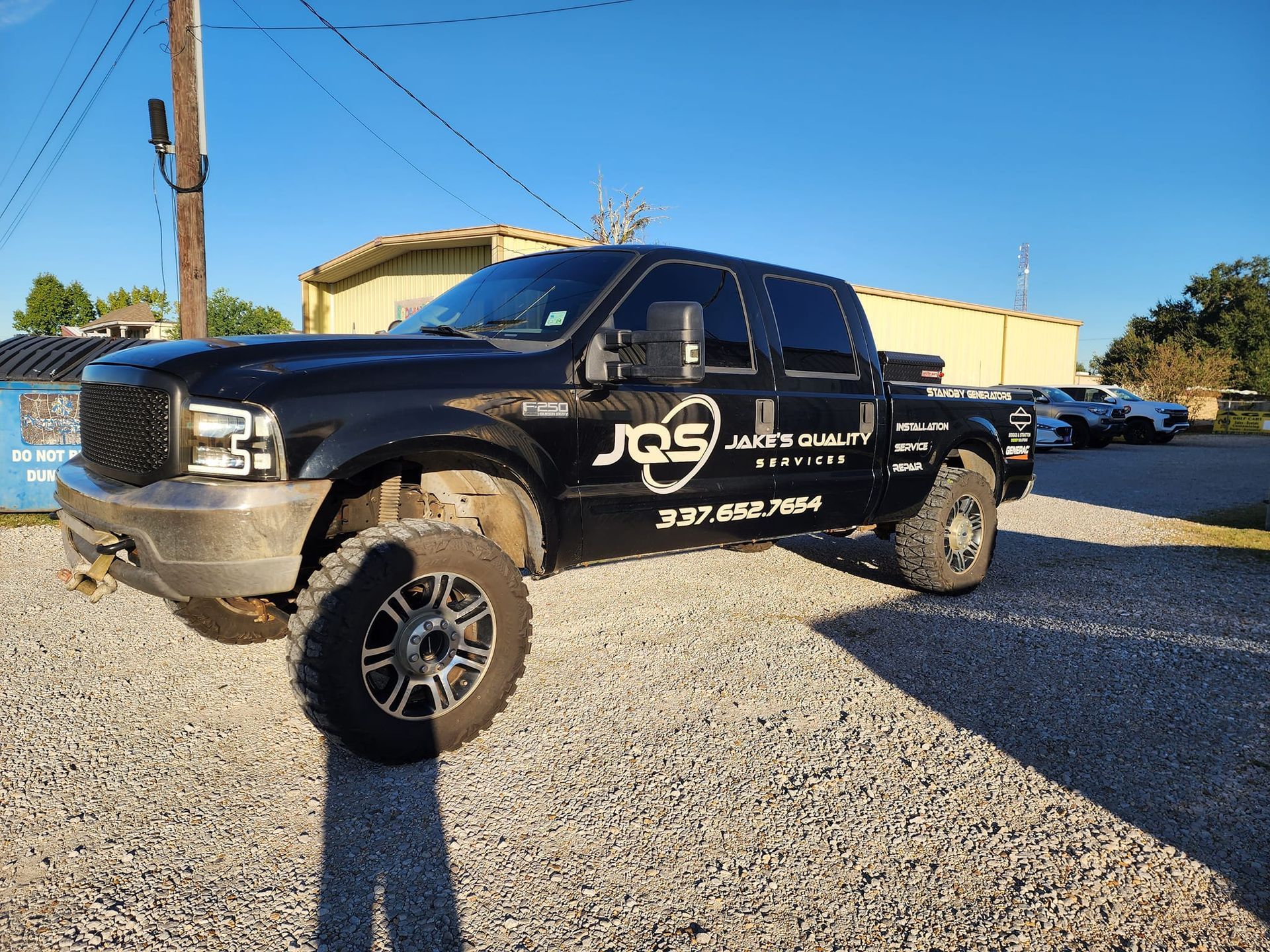 Black pickup truck with large off-road tires parked on gravel beside a tan building.