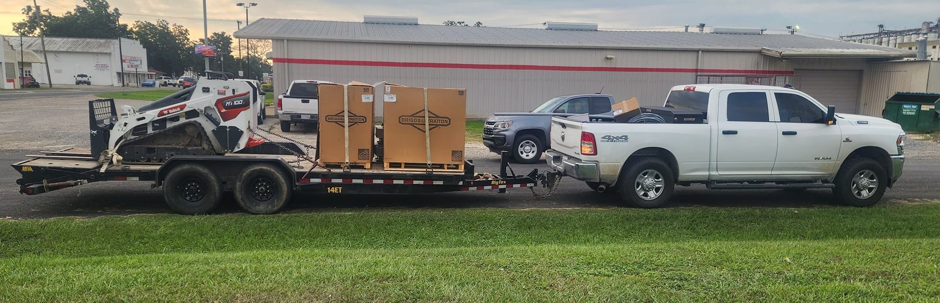 Two white pickup trucks hitched to a trailer carrying machinery beside a grassy roadside.