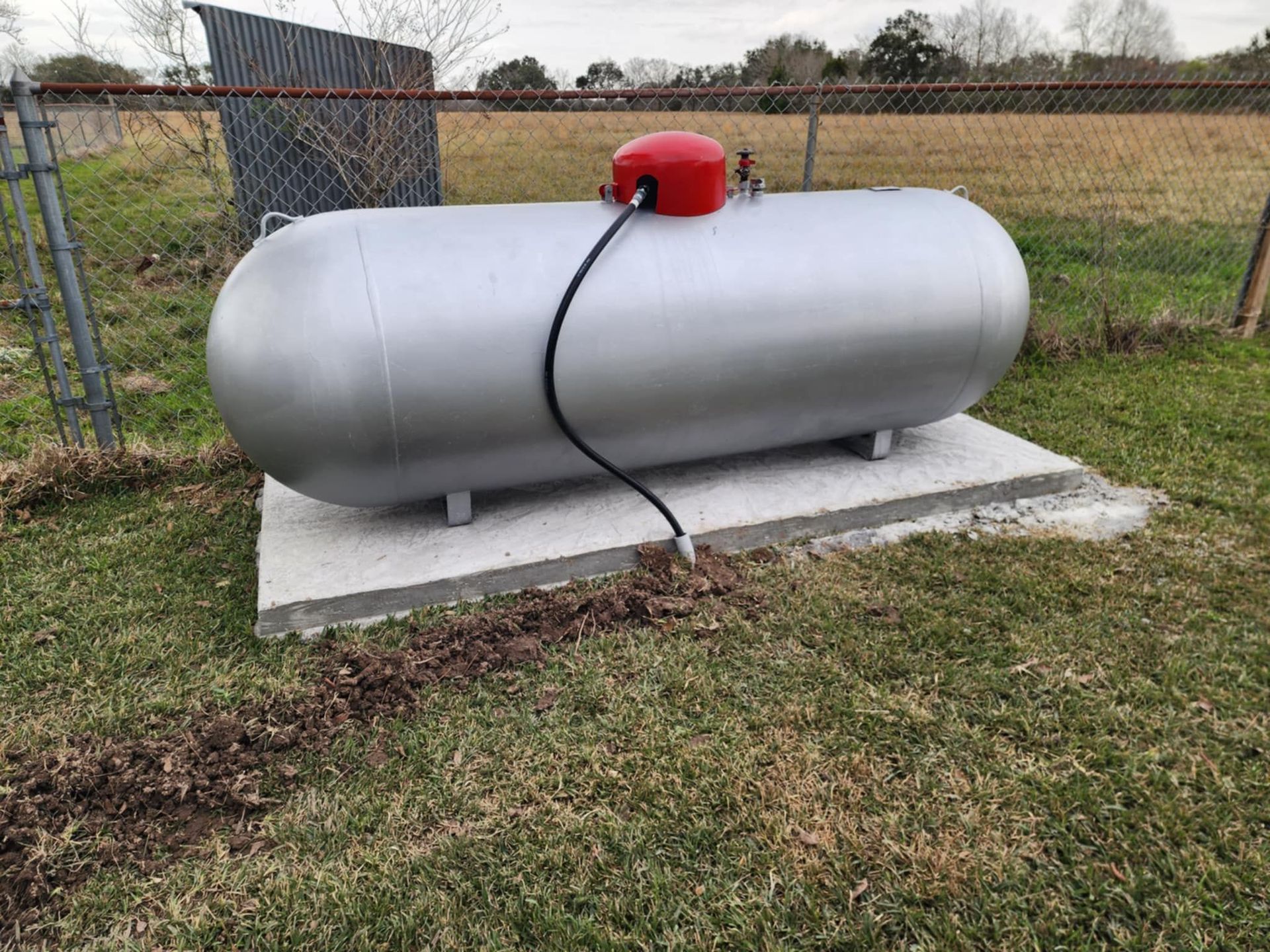 Silver propane tank on a concrete pad beside a chain-link fence, with a red valve cover and hose.
