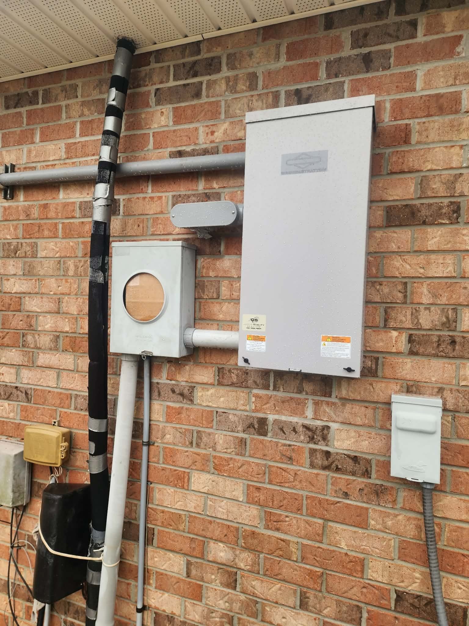 Electric meter and white utility box mounted on a brick wall with exposed pipes and cables.