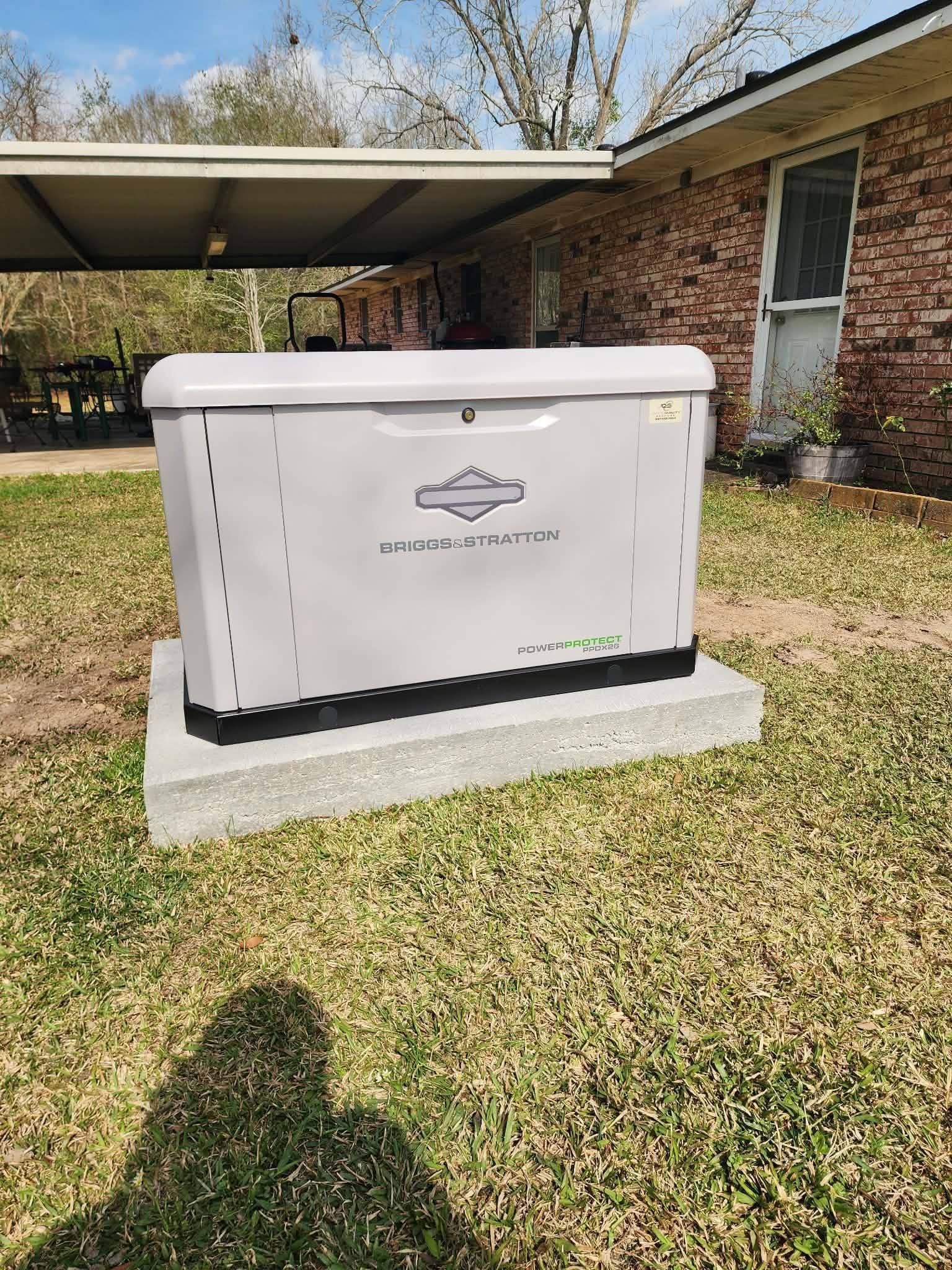 White Generac standby generator on a concrete pad beside a brick house under a carport.