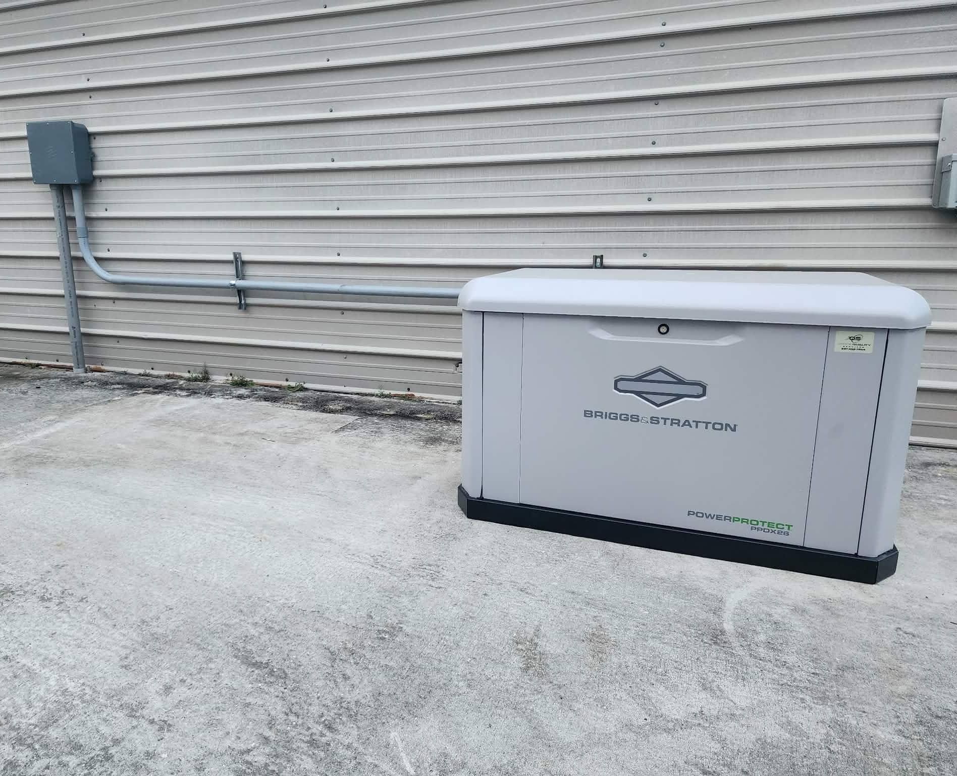 Gray standby generator beside a house, connected to the exterior wall on a concrete pad.