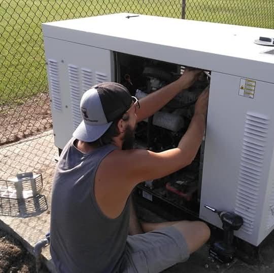 Technician kneeling beside an open outdoor generator, adjusting internal wiring near a chain-link fence.