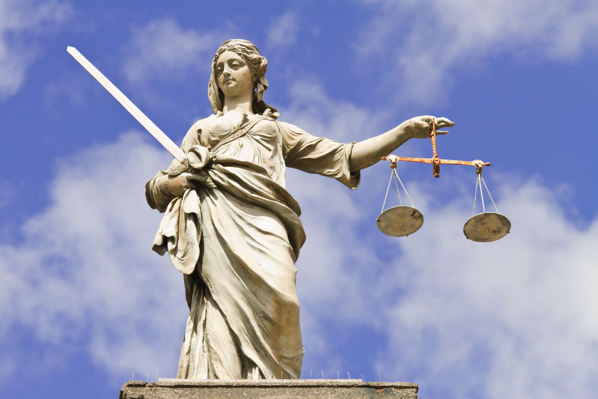 Statue of Lady Justice holding scales and a sword, standing against a cloudy blue sky.