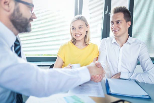 A man shaking hands with a smiling couple at a table in an office. The woman wears yellow, the man in white.