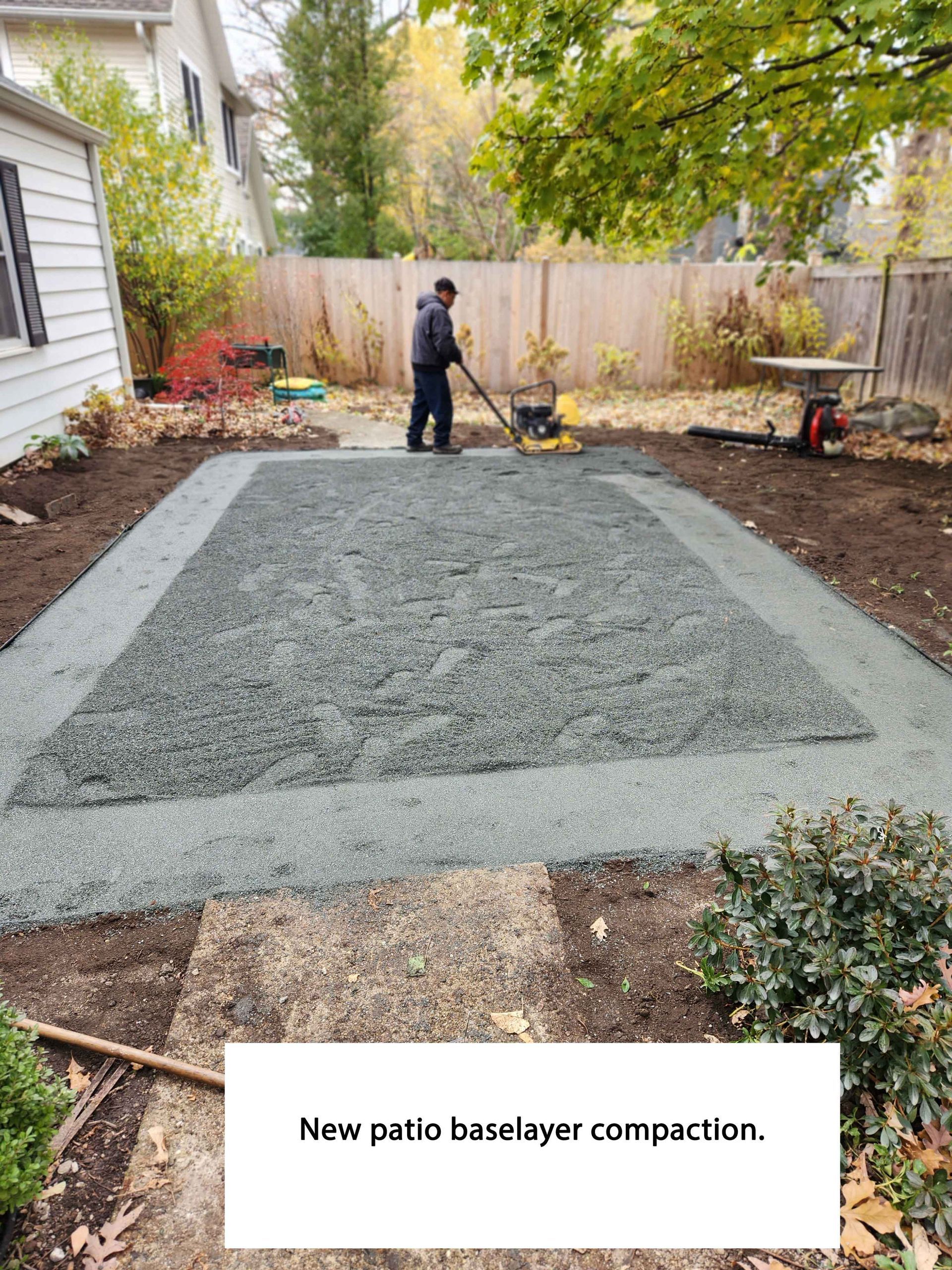 Man compacting gravel base layer for a new patio in a backyard with a house, fence, and plants.