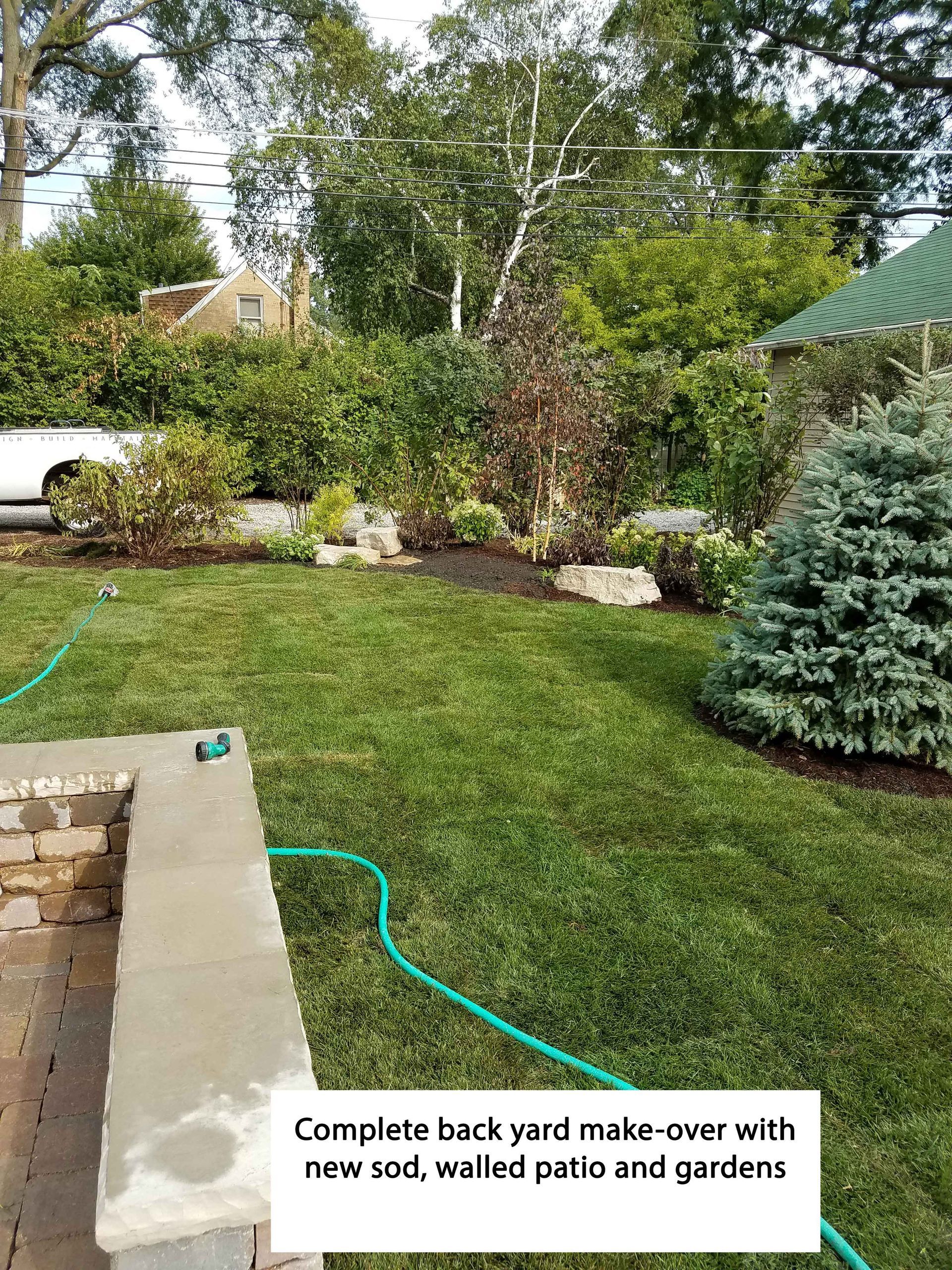 Backyard with new sod, walled patio, gardens. Green grass, trees, and a hose.
