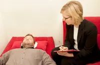 Man lying on red chaise, talking to therapist seated beside him, taking notes.