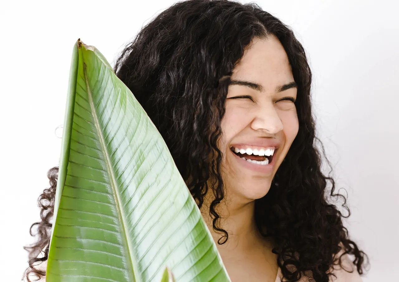 Woman laughing, holding a large green leaf, against a white background.
