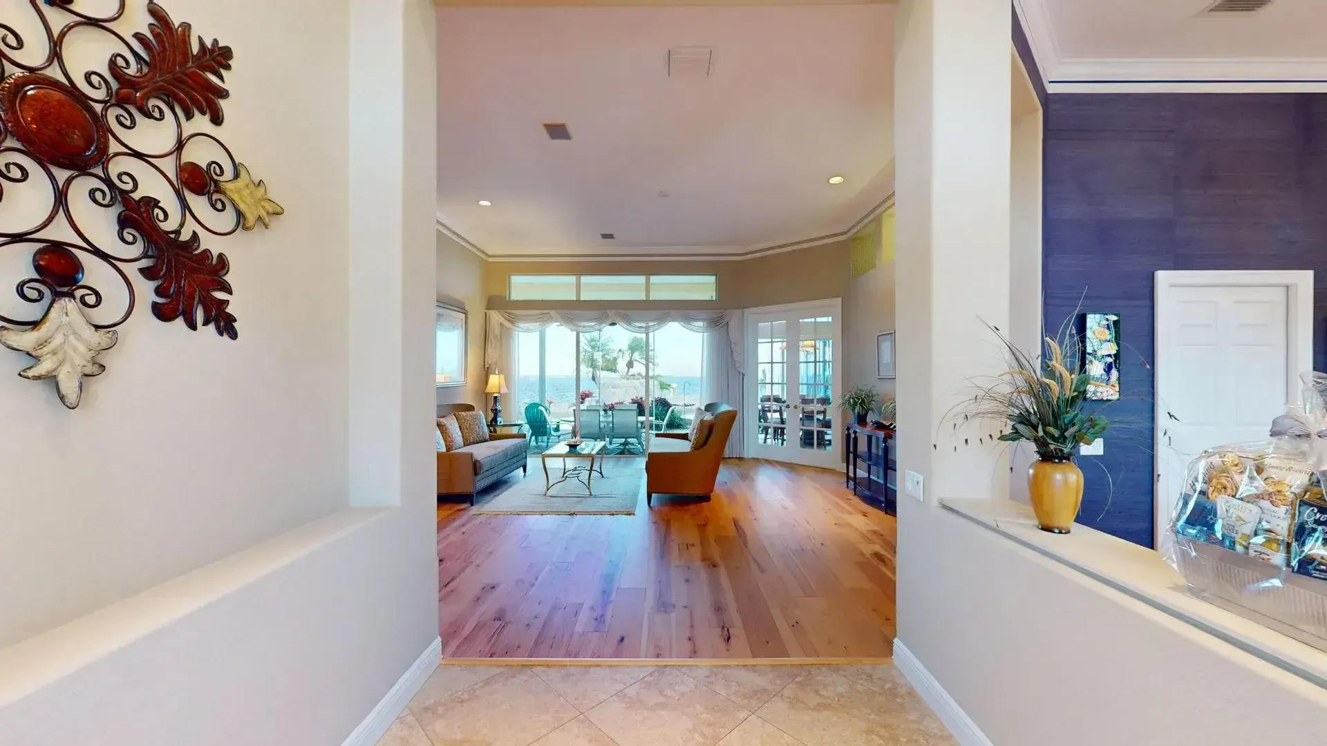Spacious living room with light hardwood floors, high ceilings, a tan sofa and armchair, and large glass sliding doors that open to a covered patio with a waterfront view.