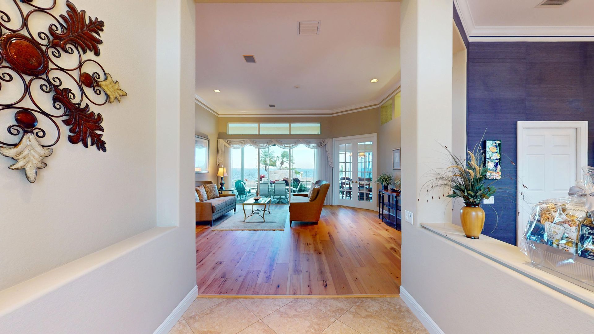 Spacious living room with light hardwood floors, high ceilings, a tan sofa and armchair, and large glass sliding doors that open to a covered patio with a waterfront view.