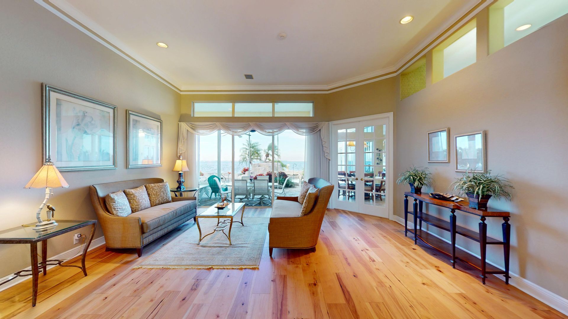 Bright, elegantly decorated living room featuring a neutral color palette, twin framed floral artwork over a classic sofa, warm accent lighting, and elegant window valences over glass doors.