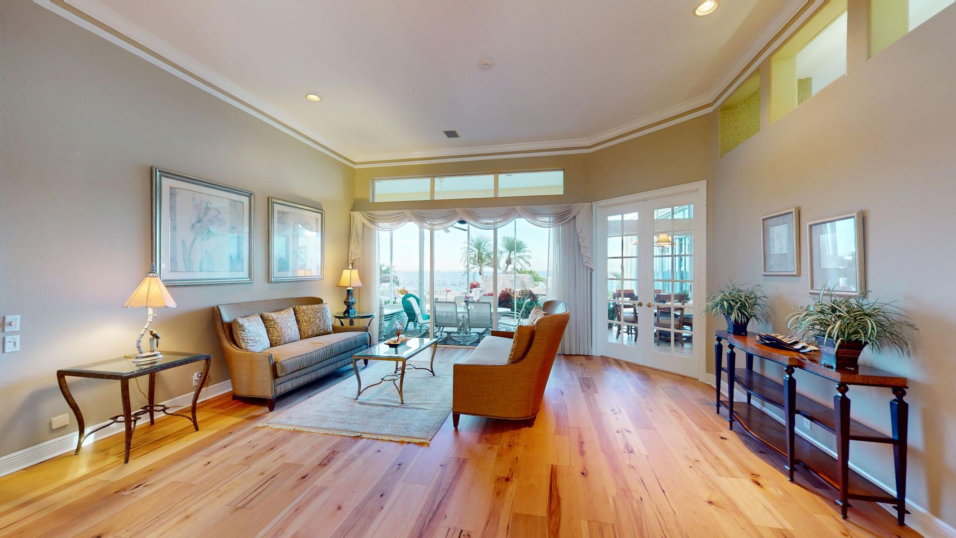 A brightly lit, upscale living room with hardwood floors, comfortable seating, and large glass doors leading to an outdoor patio with water views.