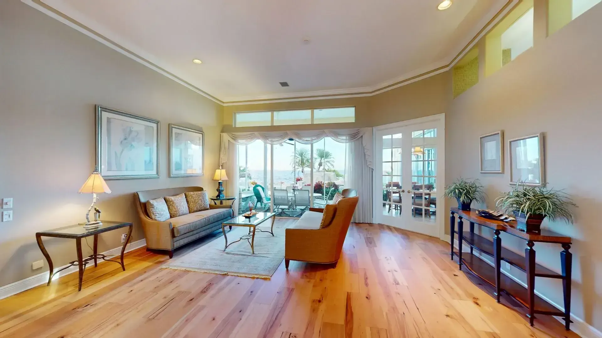 A brightly lit, upscale living room with hardwood floors, comfortable seating, and large glass doors leading to an outdoor patio with water views.