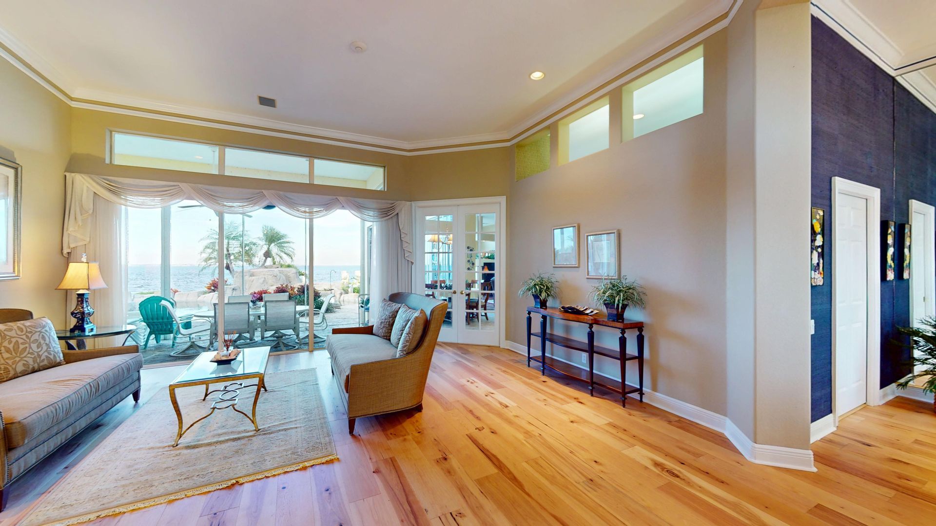 Spacious living room with light hardwood floors, high ceilings, a tan sofa and armchair, and large glass sliding doors that open to a covered patio with a waterfront view.