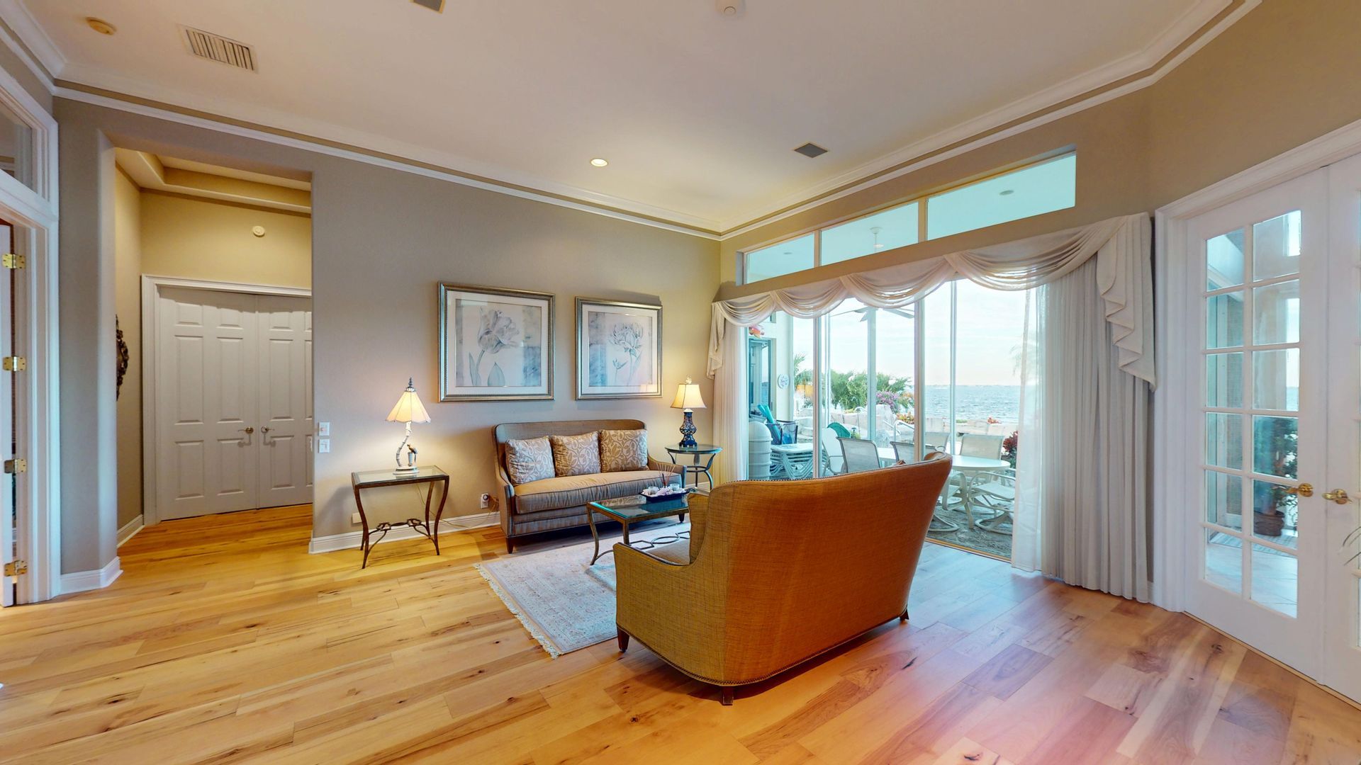 A brightly lit, upscale living room with hardwood floors, comfortable seating, and large glass doors leading to an outdoor patio with water views.
