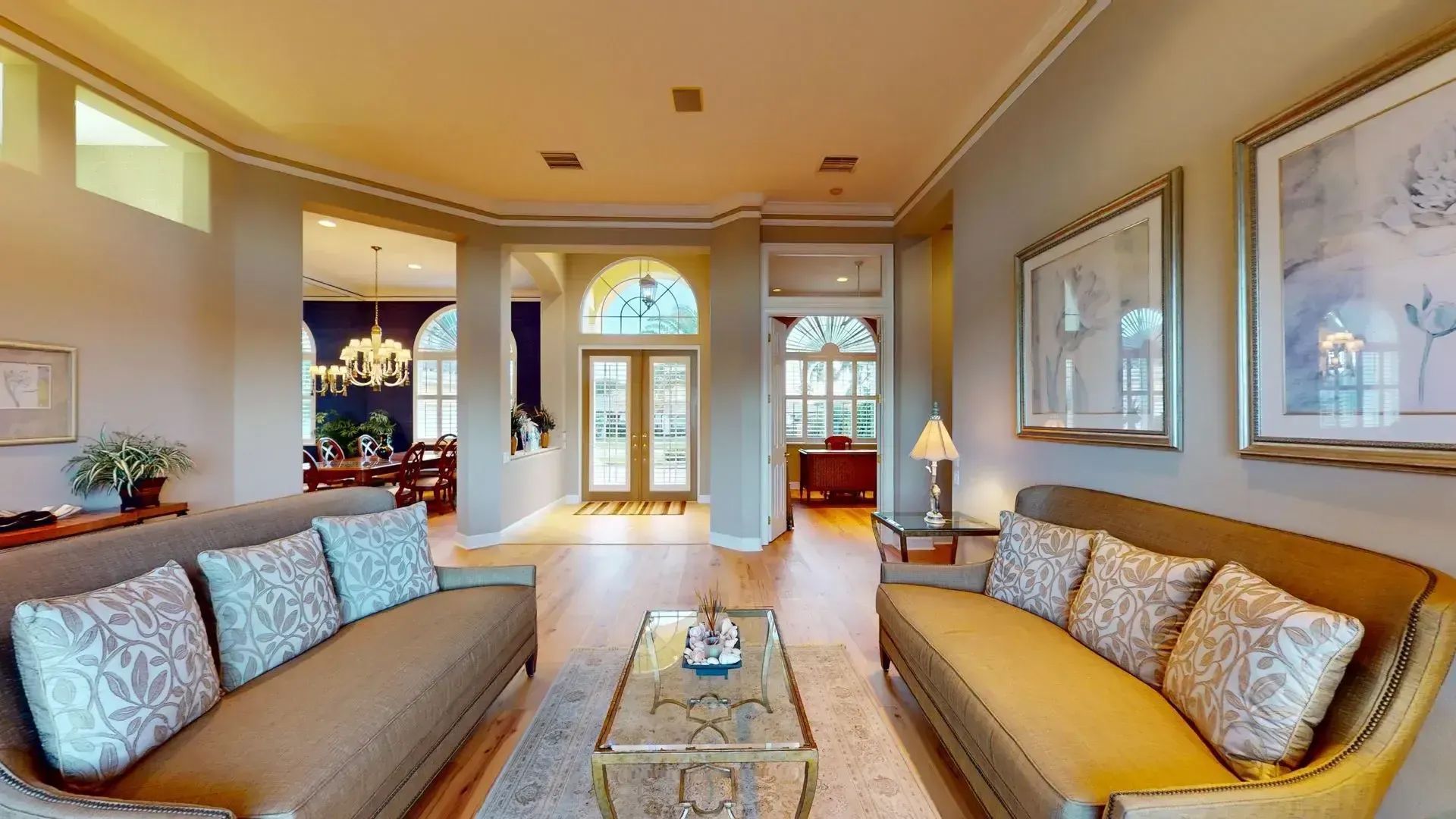 Spacious living room with light hardwood floors, high ceilings, a tan sofa and armchair, and large glass sliding doors that open to a covered patio with a waterfront view.